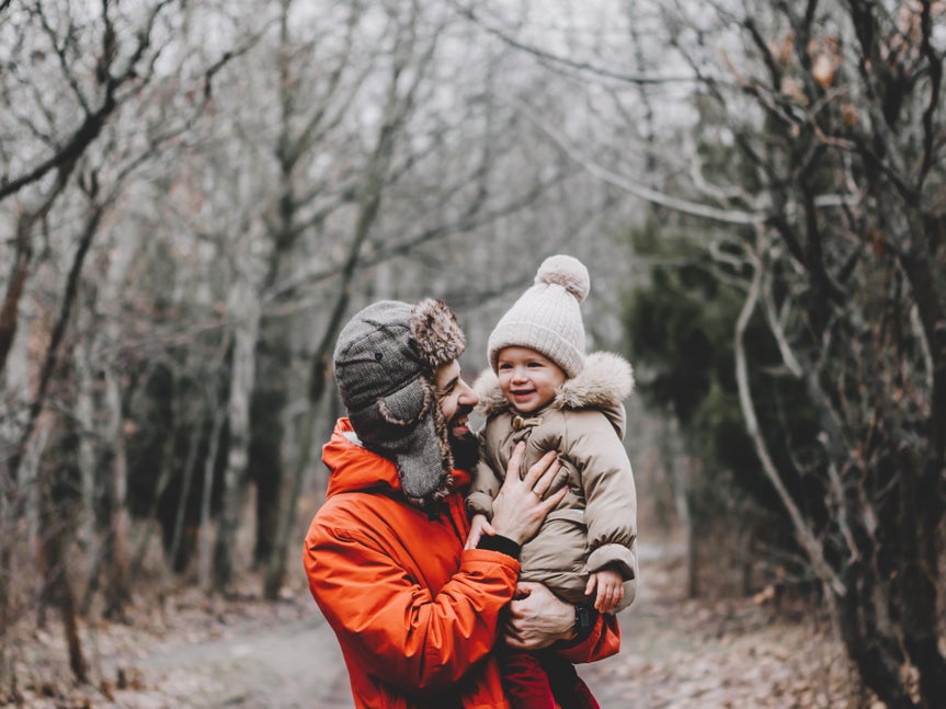 A man in an orange jacket holding a smiling baby dressed in a warm outfit, standing on a path in a leafless forest during autumn or winter