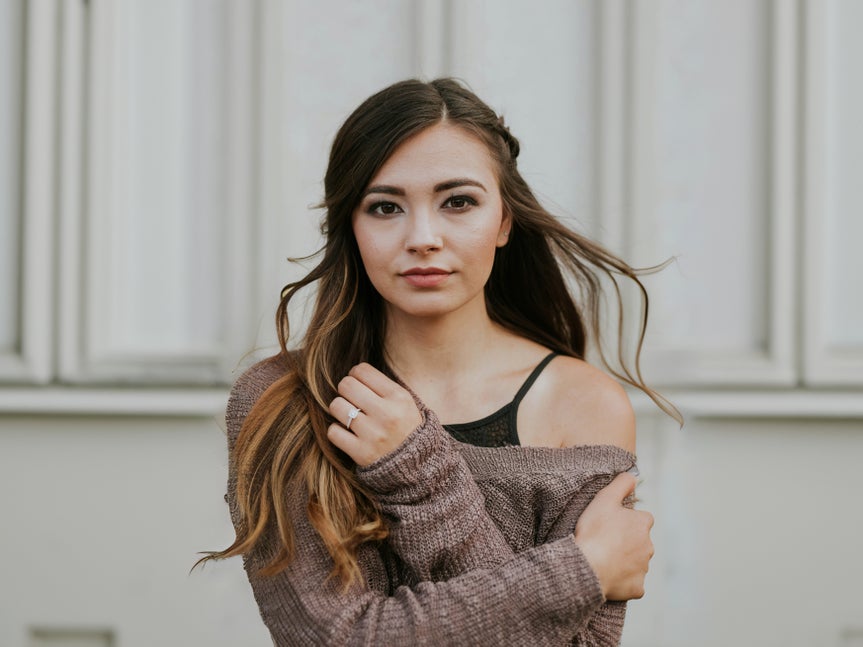 A young woman with long brown hair wearing a sweater, standing in front of a neutral background.