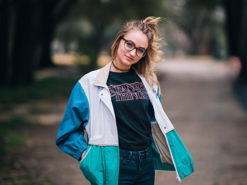 A young woman in a teal and white jacket posing outdoors with a blurred background.